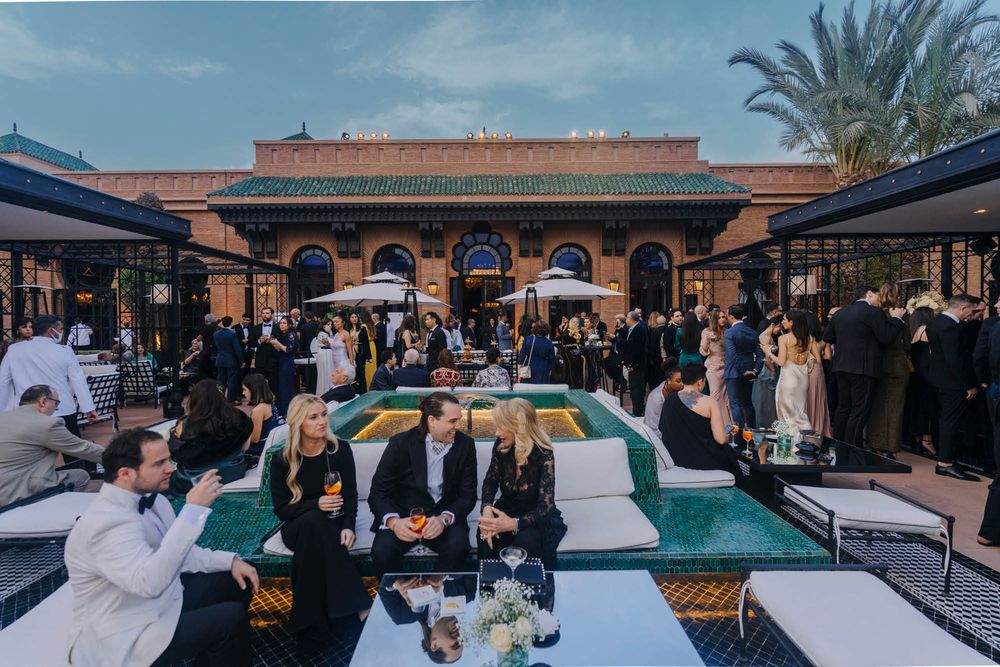 A crowded outdoor venue with guests gathered around tables under a Spanish-style architectural facade at dusk.