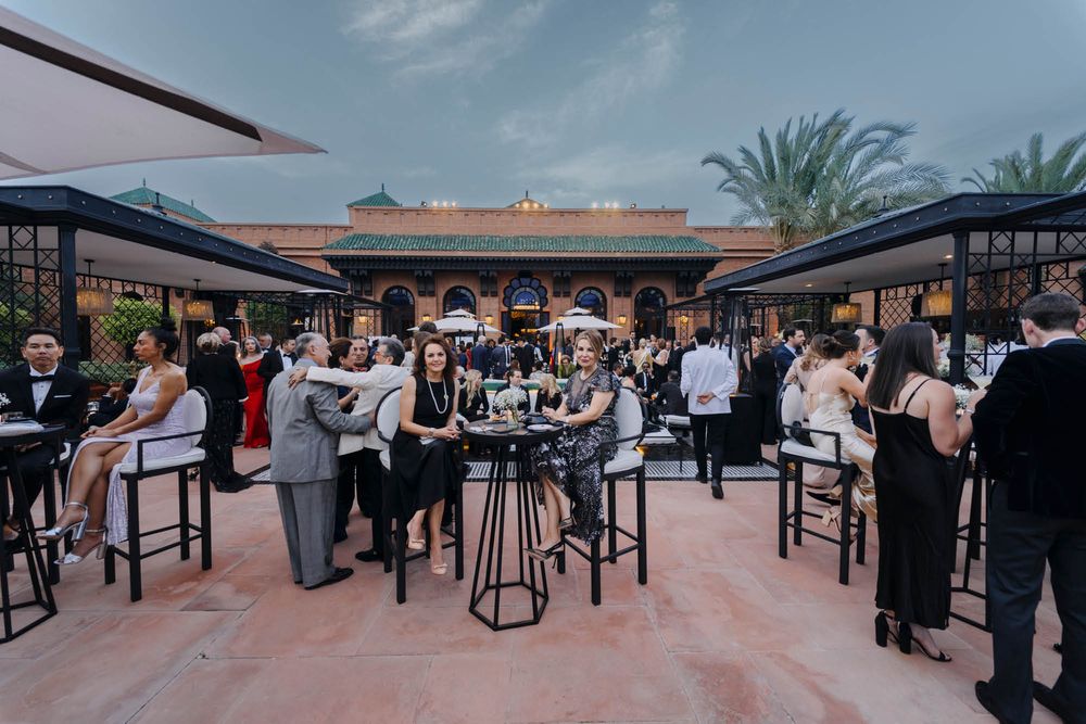 Guests mingle and socialize at an outdoor patio cocktail event with high-top tables under umbrellas at dusk.