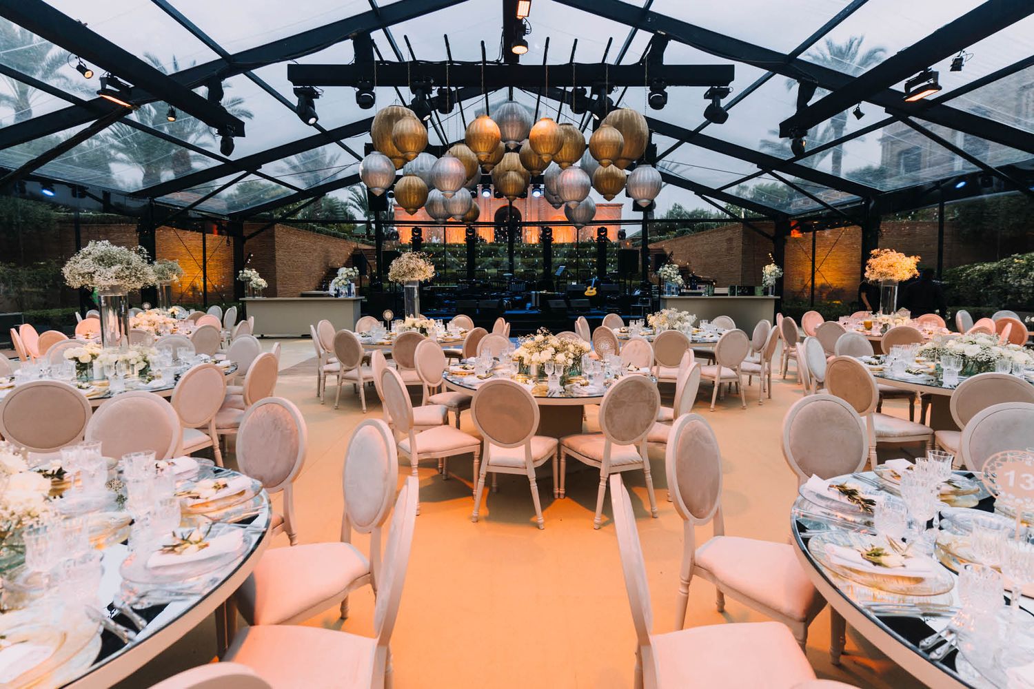 Elegant wedding reception venue with glass ceiling, pink chairs, and round tables set for dinner under hanging lantern decorations.