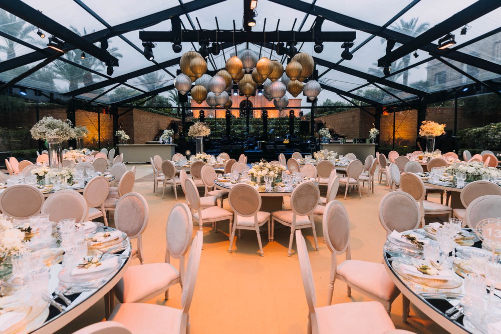 Elegant wedding reception venue with glass ceiling, pink chairs, and round tables set for dinner under hanging lantern decorations.
