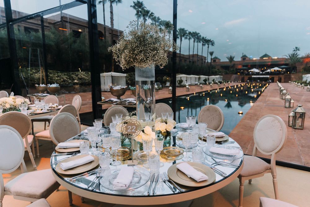 Elegant outdoor wedding reception setup with round tables and white chairs arranged beside a reflective pool under palm trees.