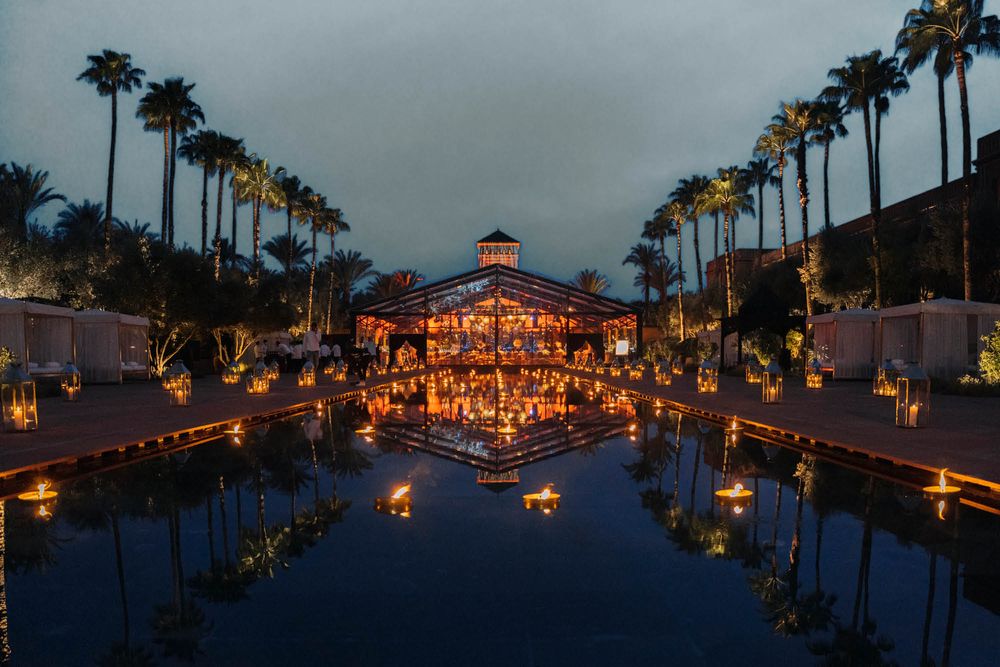 Palm trees line a reflection pool leading to an illuminated pavilion at twilight with warm glowing lanterns creating a magical scene.