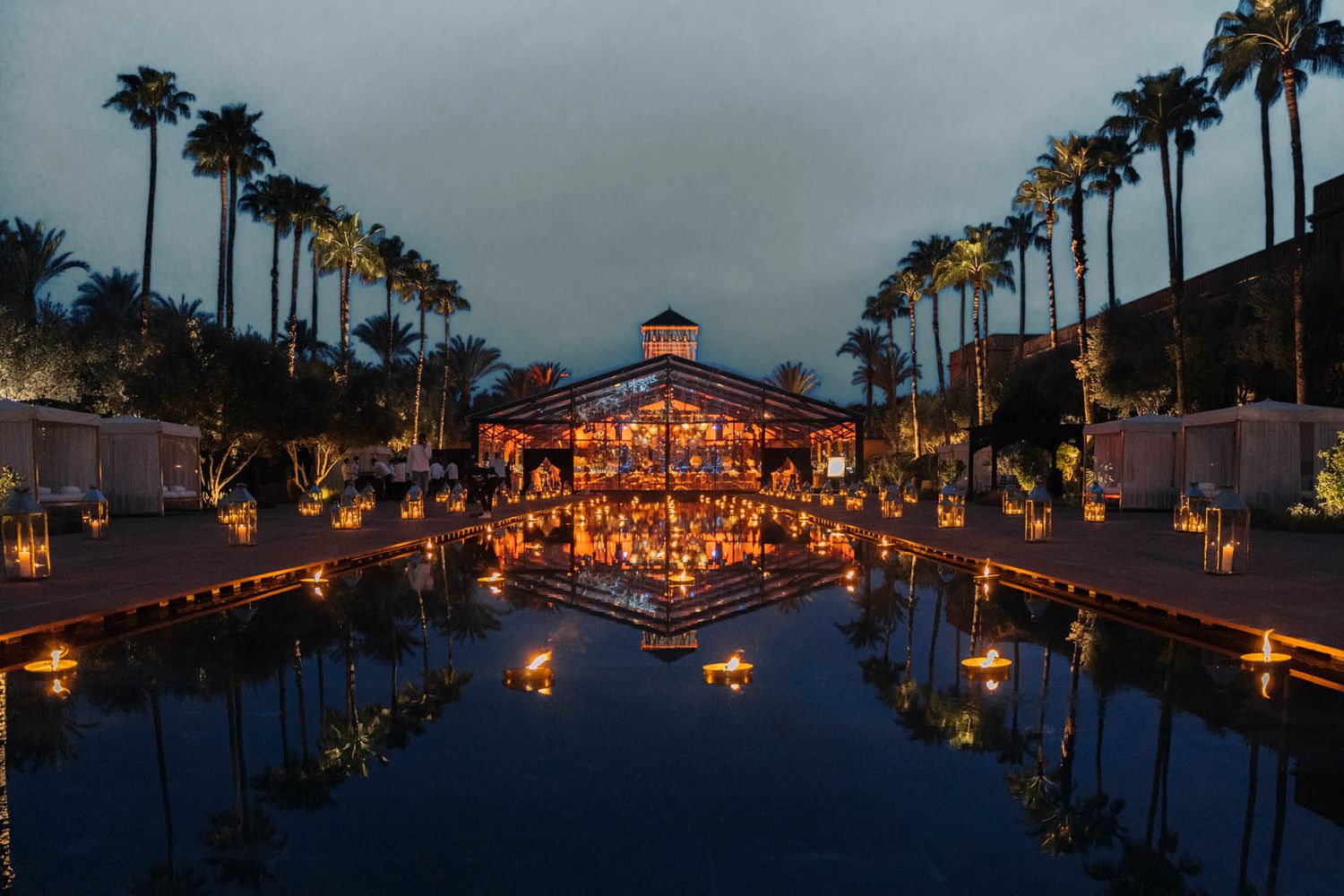 Palm trees line a reflection pool leading to an illuminated pavilion at twilight with warm glowing lanterns creating a magical scene.