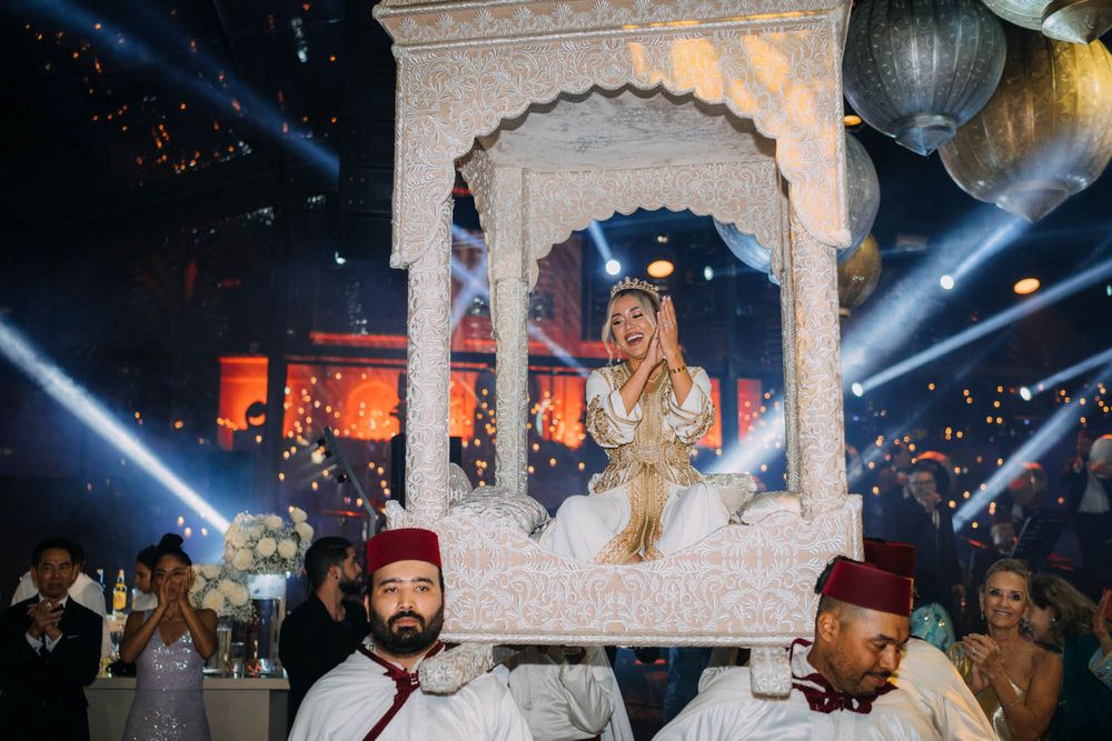 A stage performance with a decorated white ornate arch frame being carried by performers in red fez hats amid dramatic lighting.