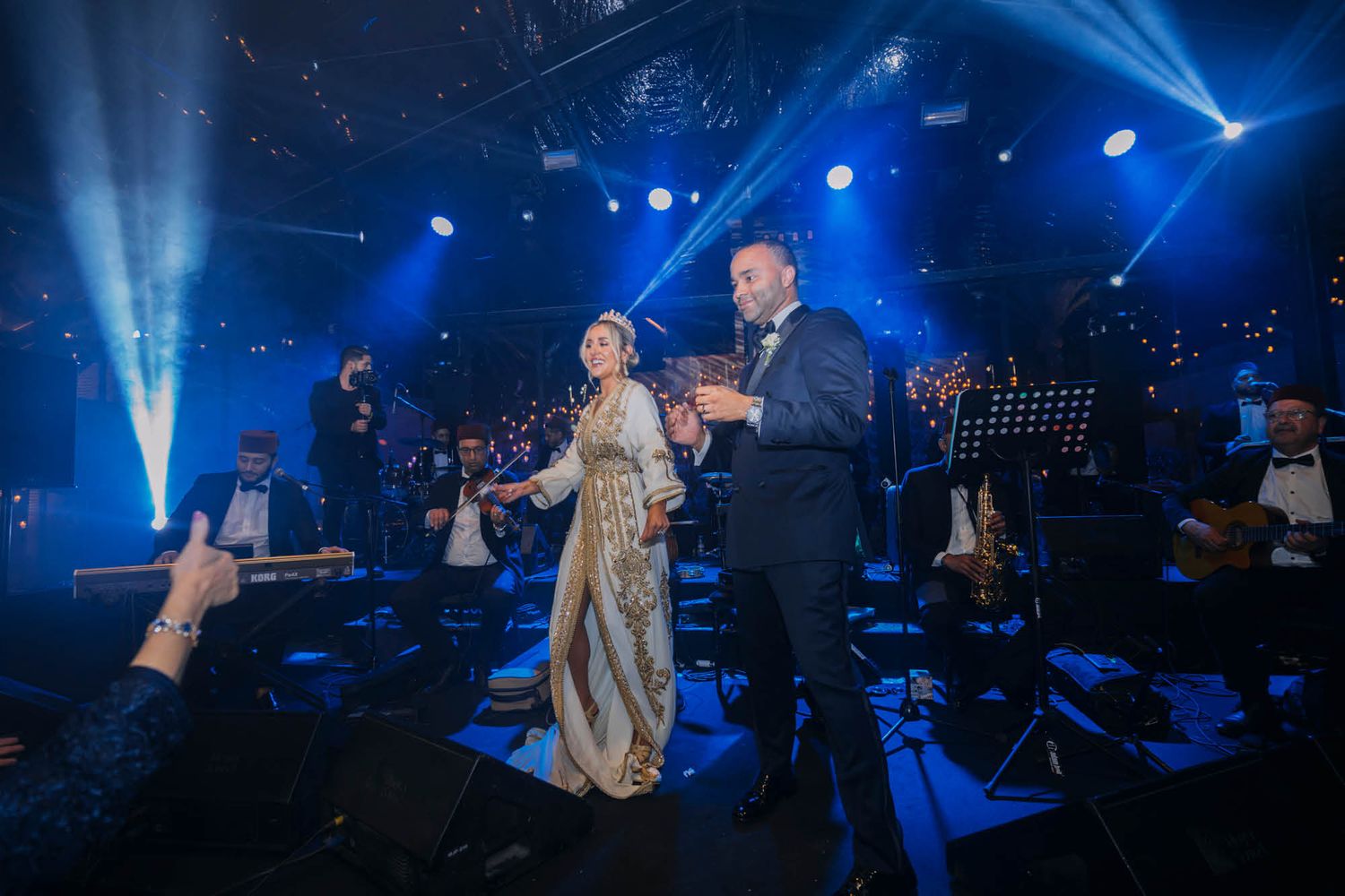 Two performers sing in elegant formal wear on a concert stage lit by dramatic blue spotlights and beams.