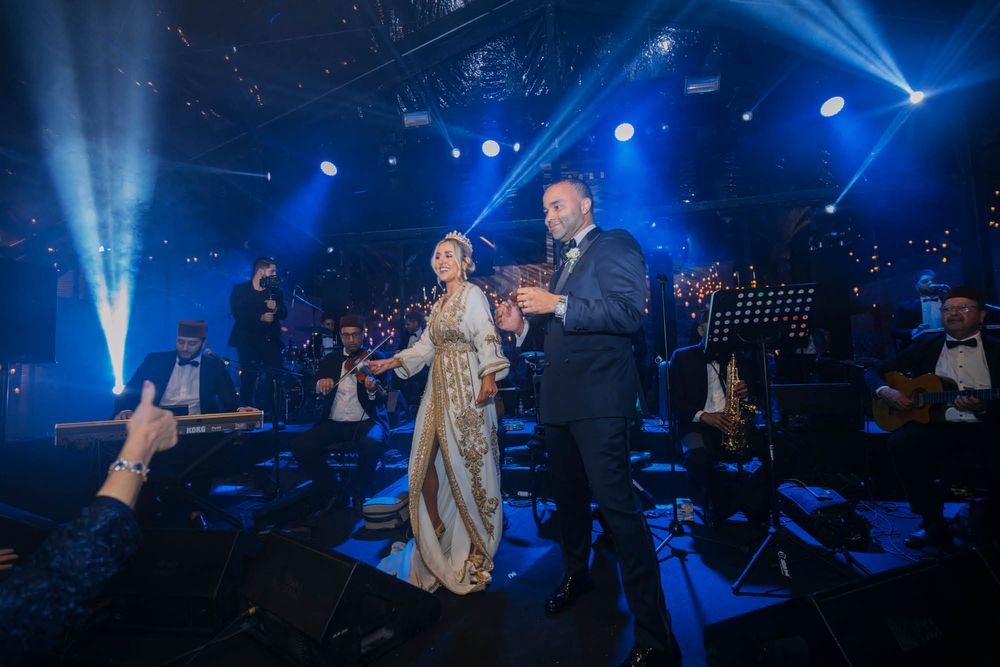Two performers sing in elegant formal wear on a concert stage lit by dramatic blue spotlights and beams.