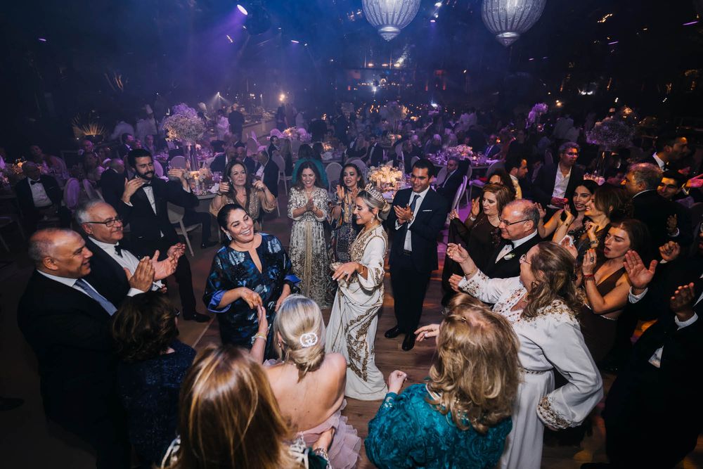 Wedding reception crowd dancing and celebrating on a dark dance floor illuminated by overhead lanterns and party lights.
