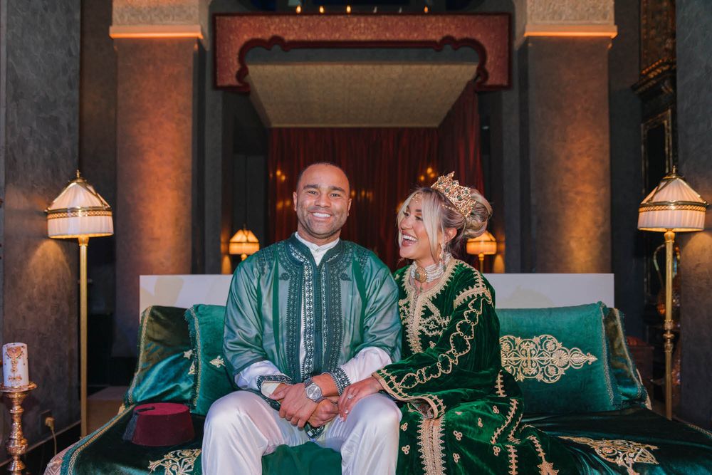 Two people sit together on green velvet couch wearing traditional Moroccan formal attire in an elegant interior setting.