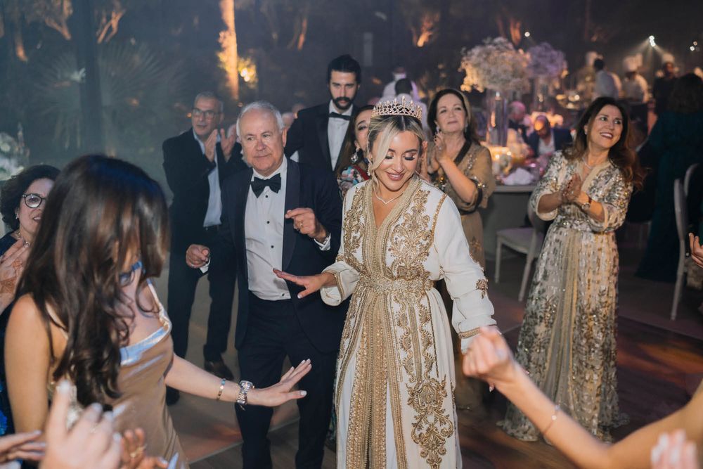Wedding guests dance and celebrate on a dimly lit dance floor with people wearing formal attire including gold dresses.