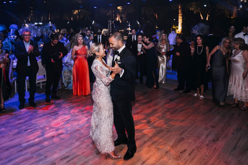 A couple shares a romantic dance on a wooden floor under colorful lights while guests watch at a formal evening event.