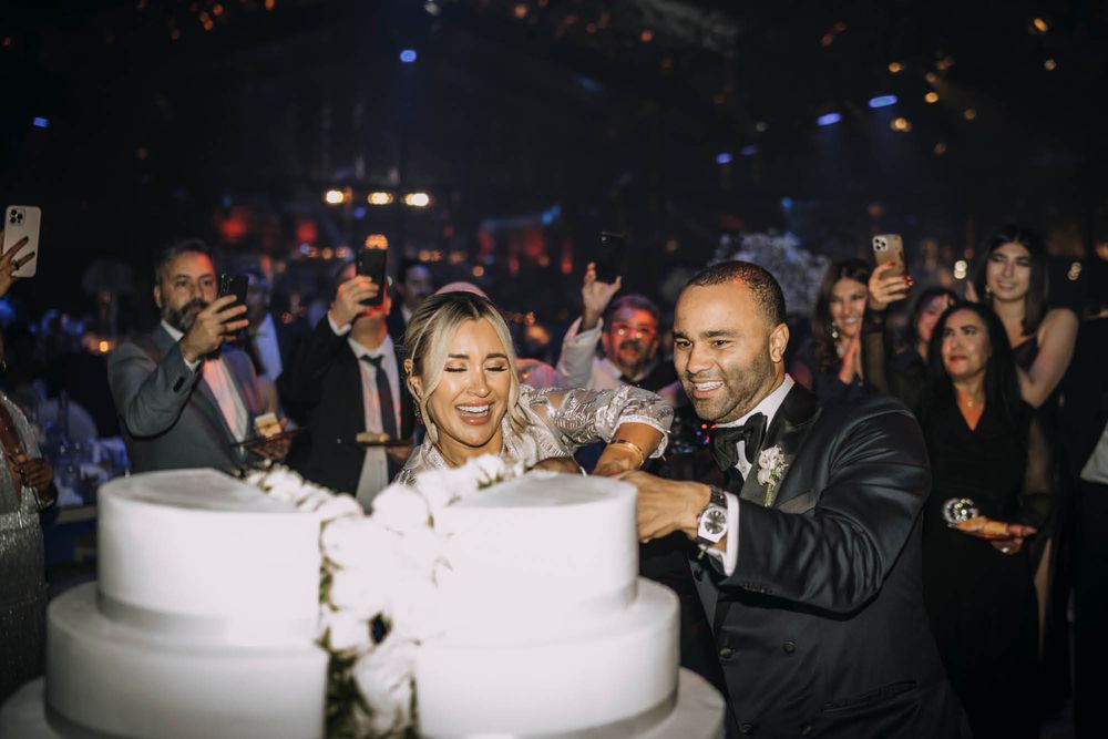 Couple celebrates at an elegant celebration cutting into a large two-tiered white wedding cake in a dimly lit venue.