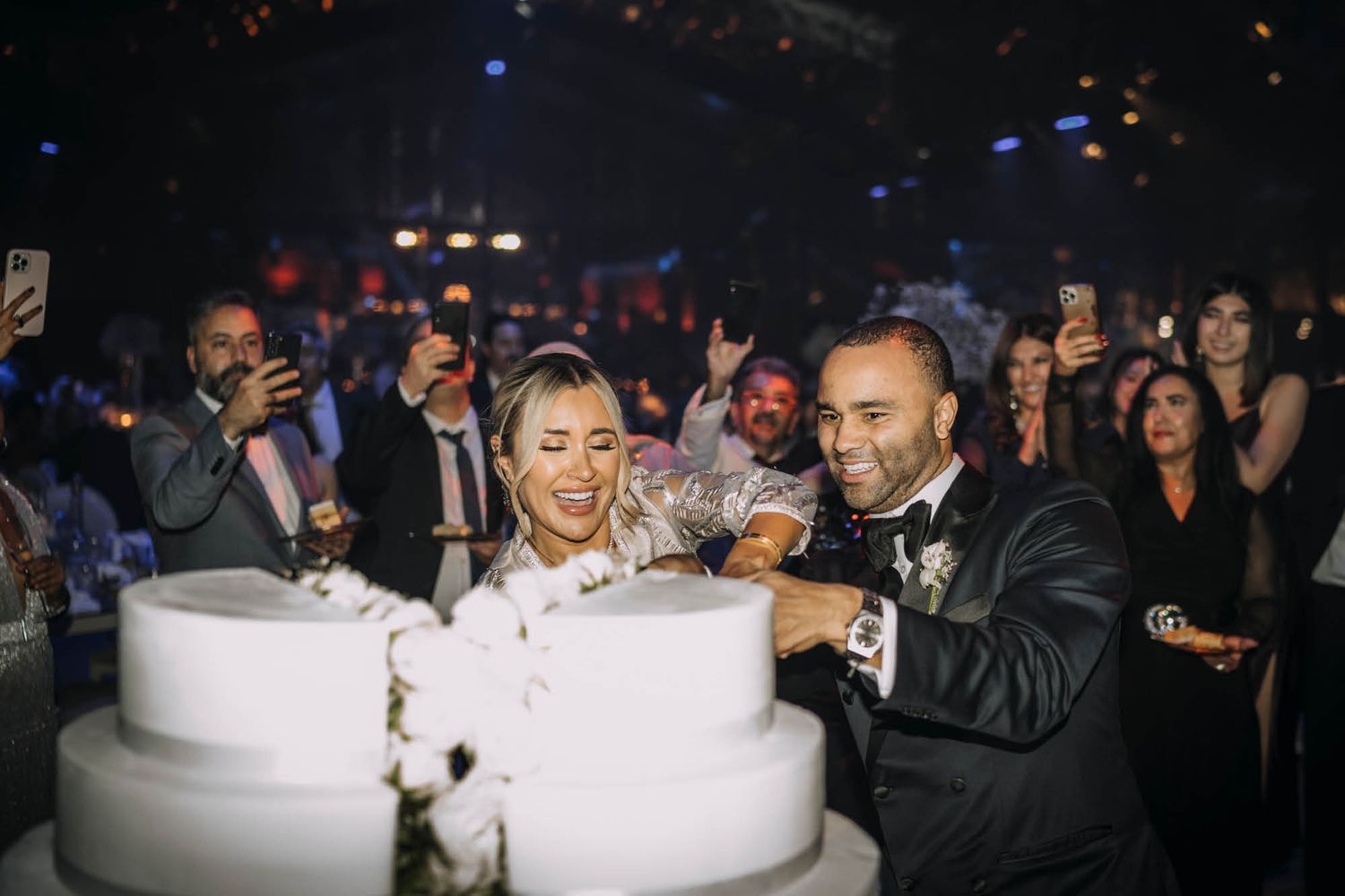 Couple celebrates at an elegant celebration cutting into a large two-tiered white wedding cake in a dimly lit venue.