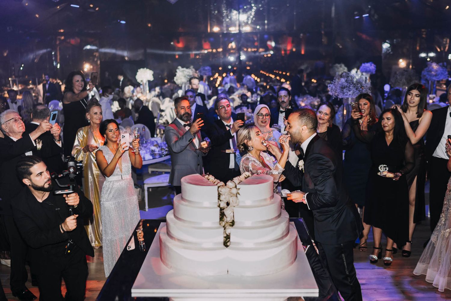 A large wedding crowd gathers around an elegant tiered white cake at a dimly lit reception venue with blue lighting.