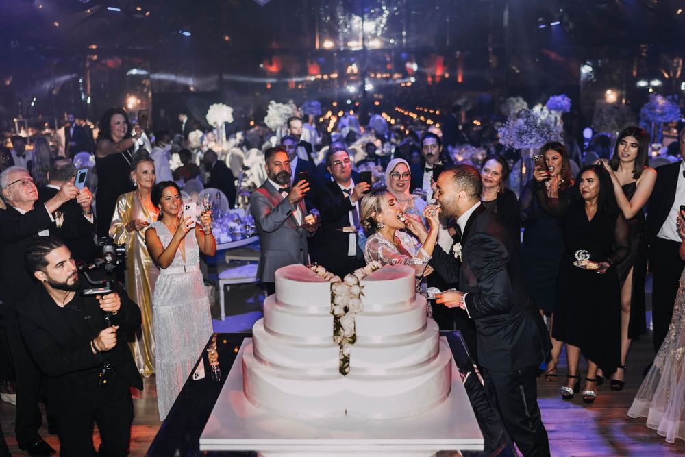 A large wedding crowd gathers around an elegant tiered white cake at a dimly lit reception venue with blue lighting.