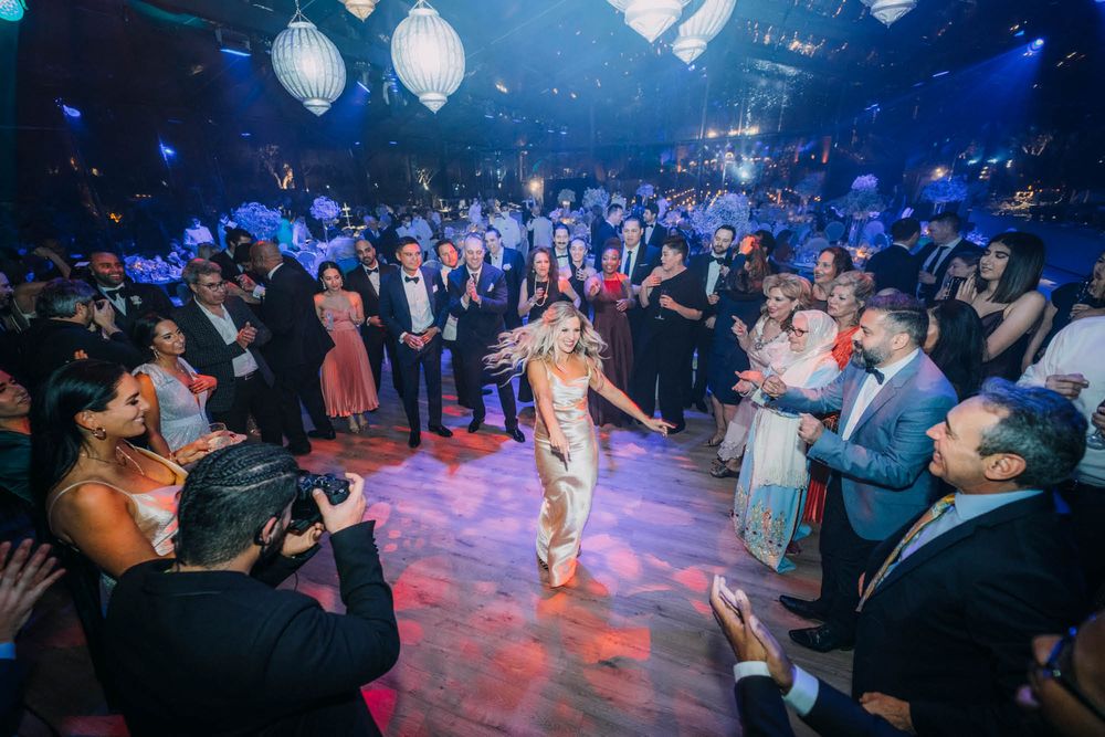 A crowded nightclub dance floor illuminated with blue and purple lights as partygoers dance and take photos.