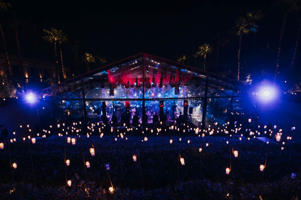 Illuminated outdoor concert venue at night with glowing lanterns scattered across the ground and palm trees in background.