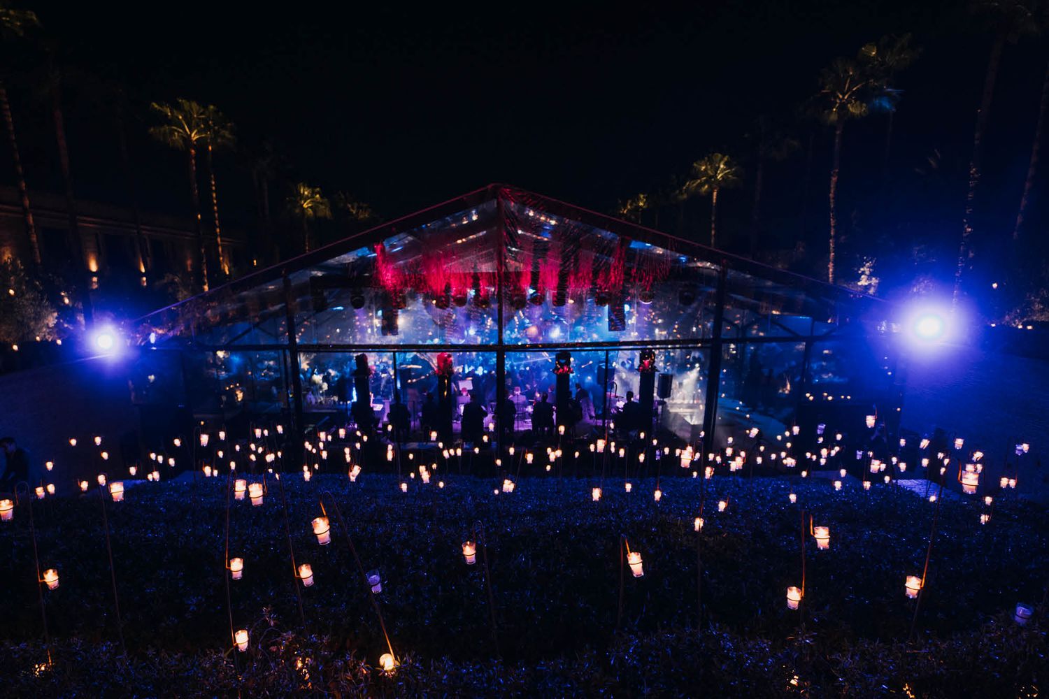 Illuminated outdoor concert venue at night with glowing lanterns scattered across the ground and palm trees in background.