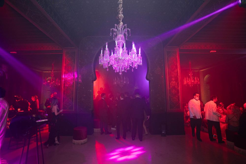 A crystal chandelier glows with purple lighting above a dance floor filled with silhouettes in a nightclub atmosphere.
