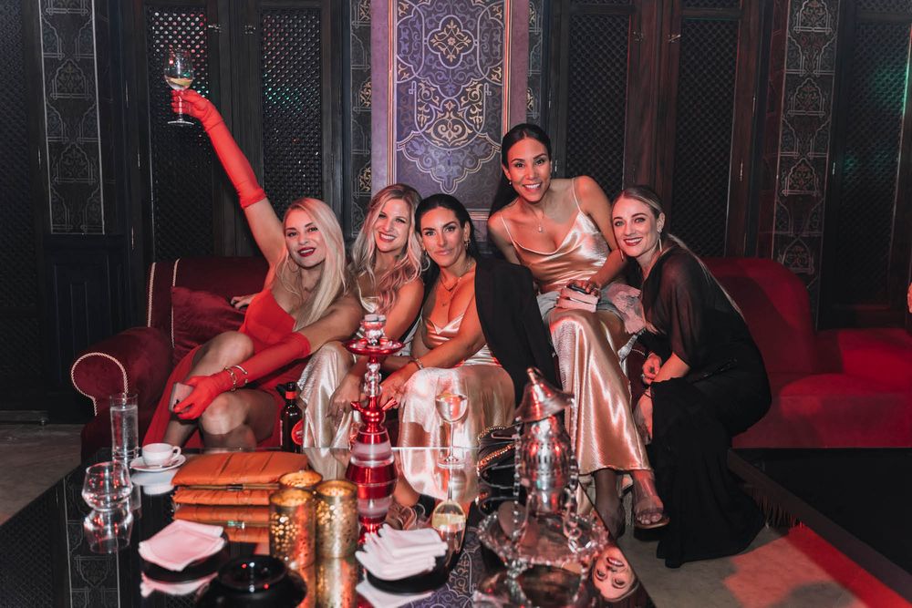 A group celebrates at a nightclub lounge with drinks and hookah pipes on the table in moody red lighting.