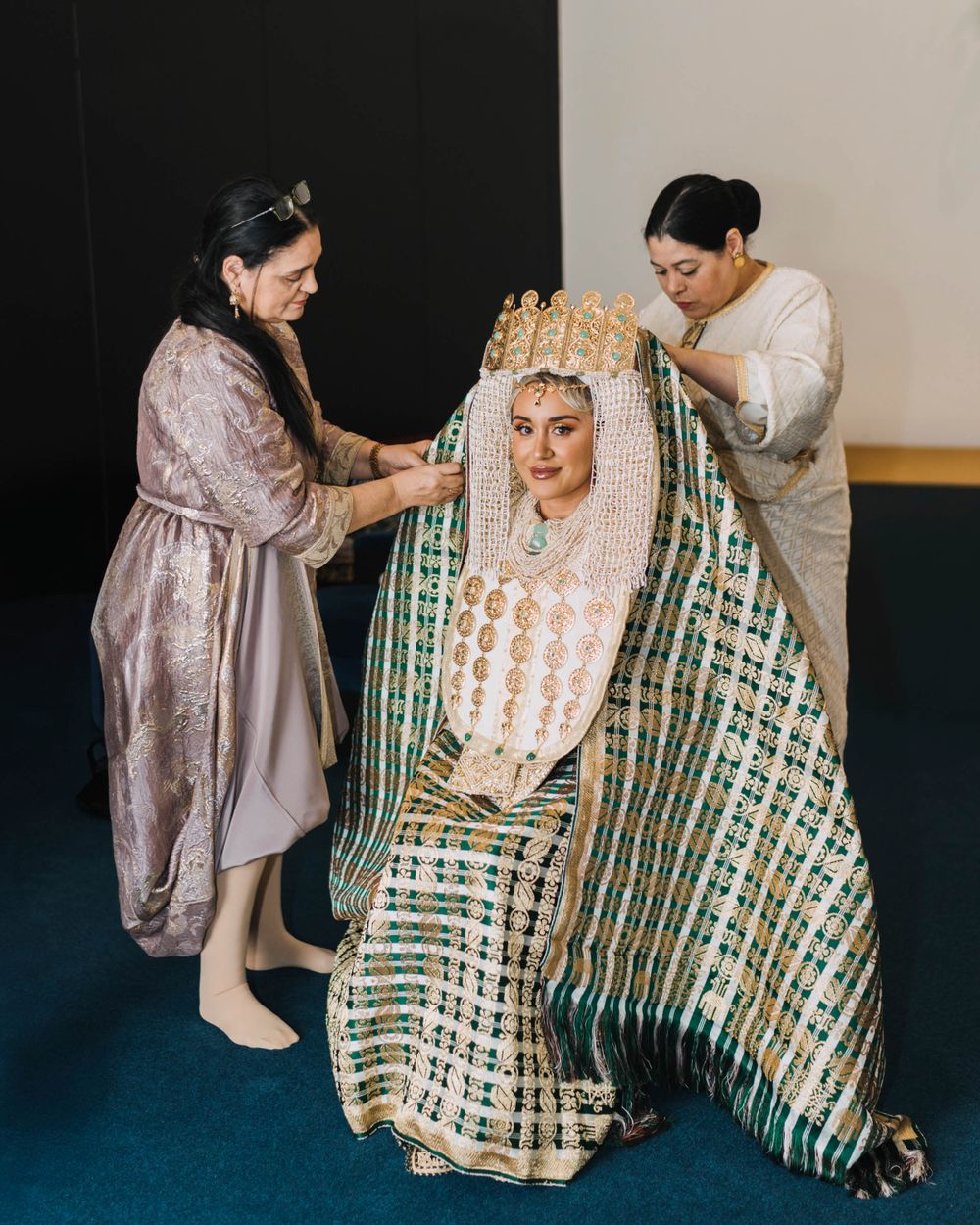 Traditional ceremonial wedding preparation showing two helpers adjusting an ornate green and gold patterned robe and headdress.