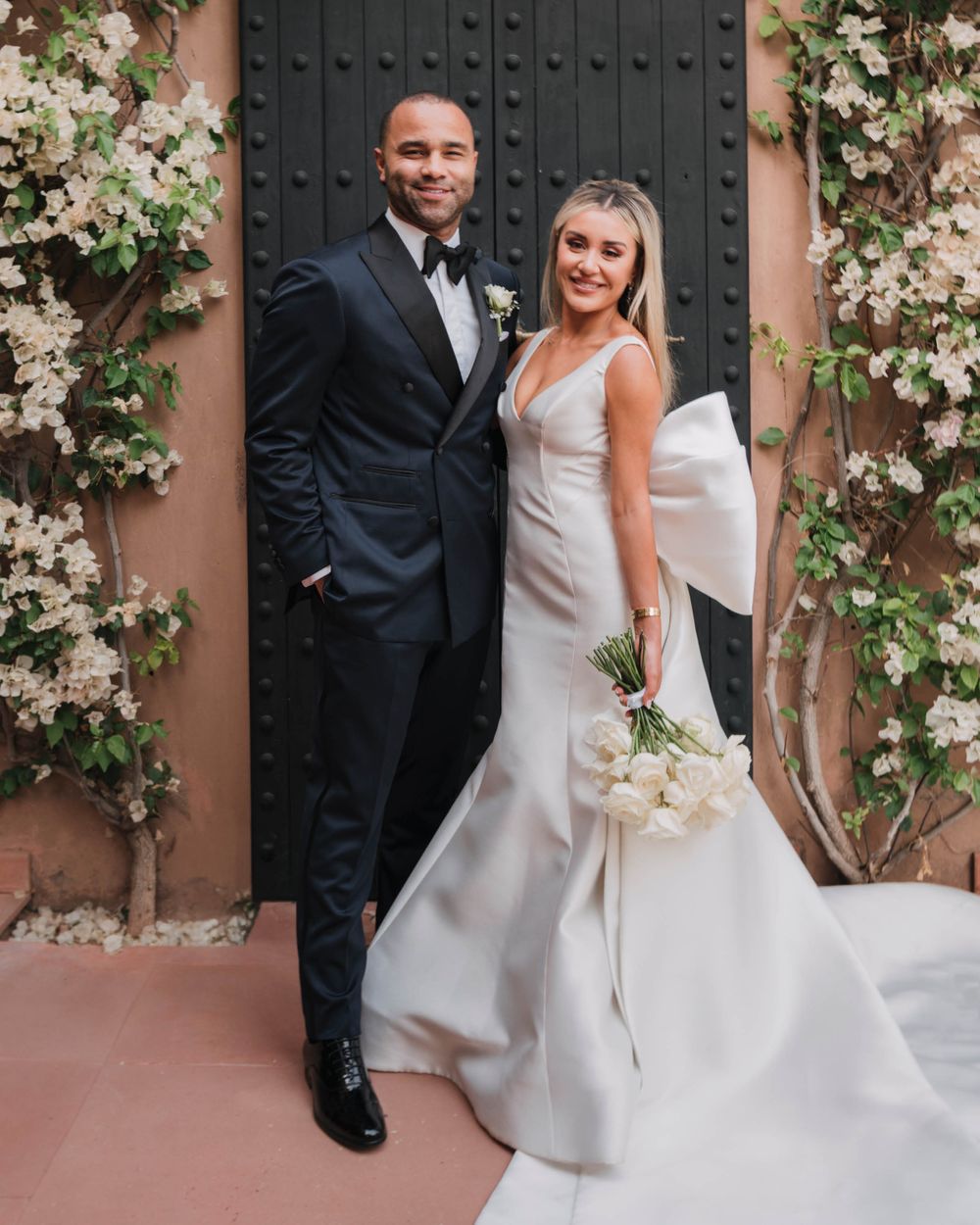 Wedding couple poses together in front of a rustic door with climbing white flowers and blossoming vines.