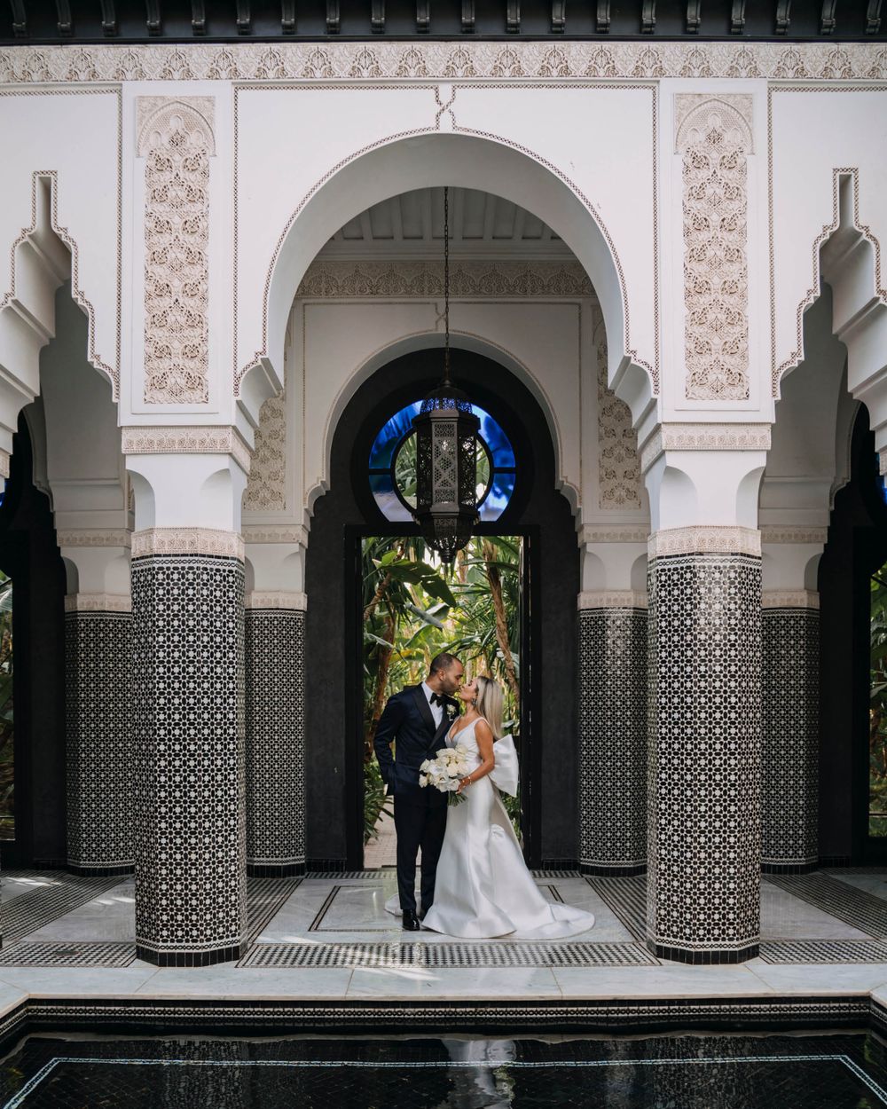 A newlywed couple stands under ornate Moroccan archways with intricate tile work and traditional Islamic architecture.