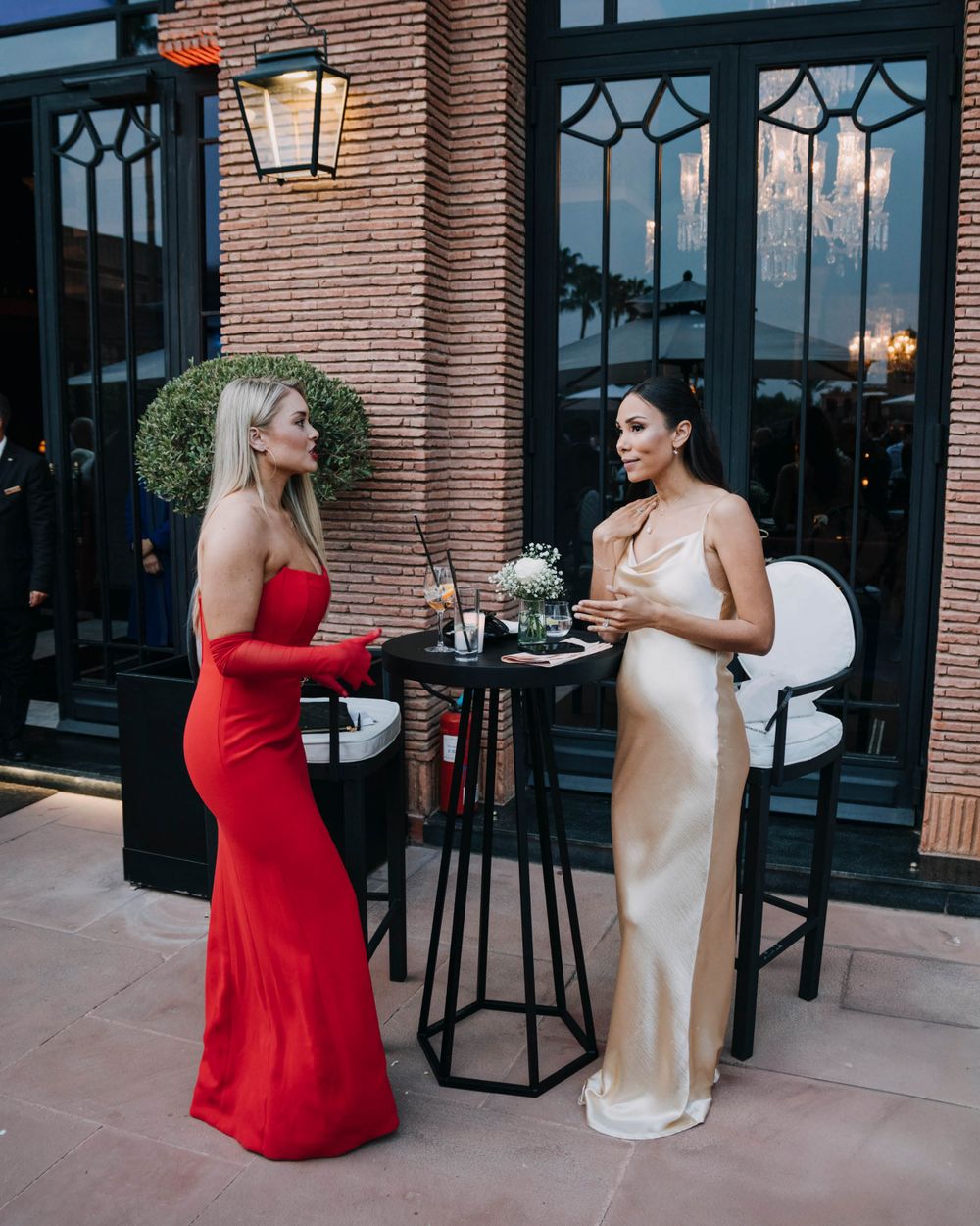 Two women in elegant evening gowns, one red and one champagne-colored, chat while standing on an outdoor patio at dusk.