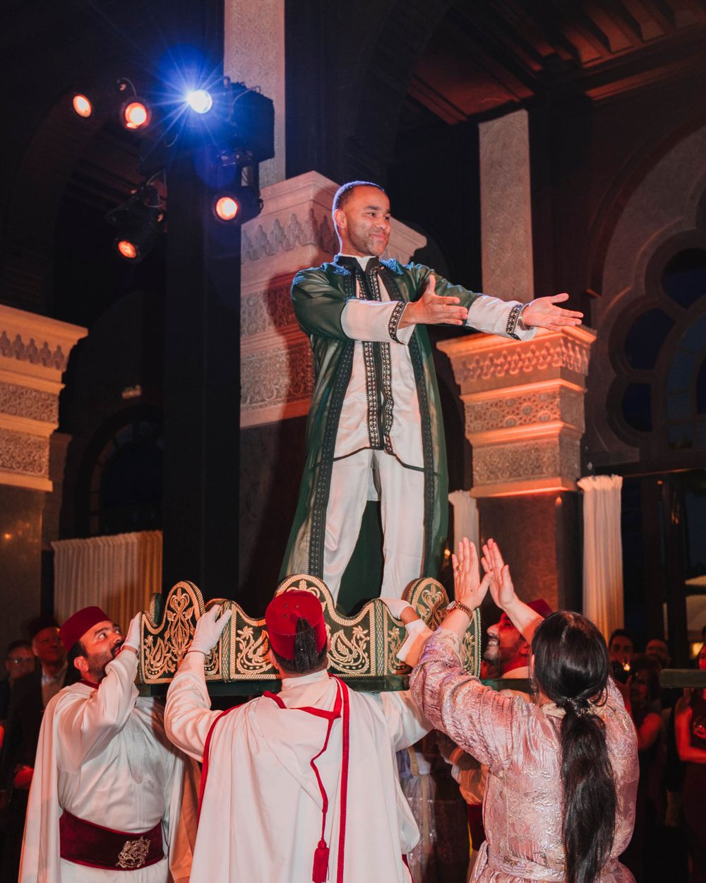 Performer in traditional costume stands elevated above crowd during vibrant cultural celebration at ornate venue.