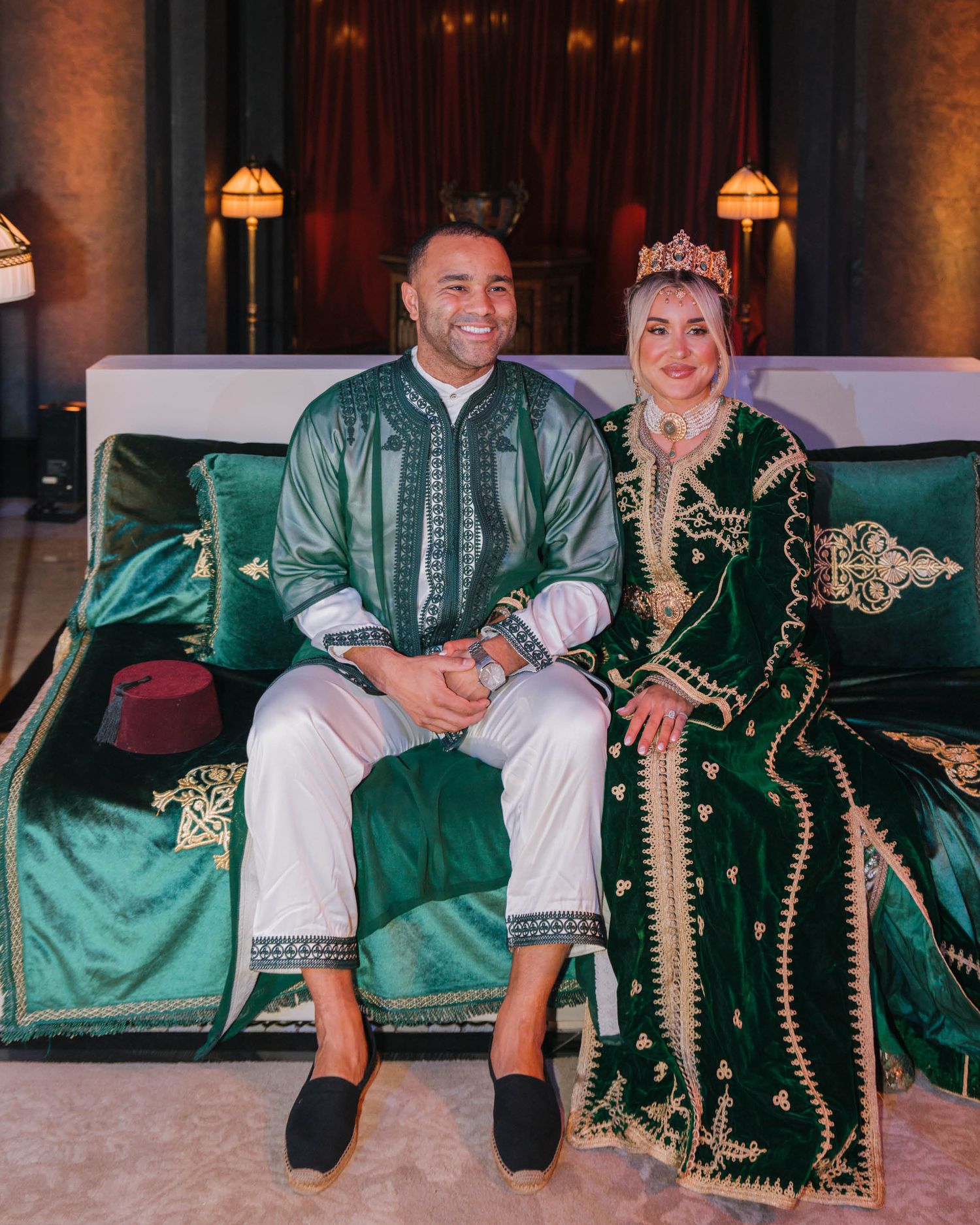 A couple in traditional Moroccan dress sits together on an emerald green velvet couch with ornate gold details.
