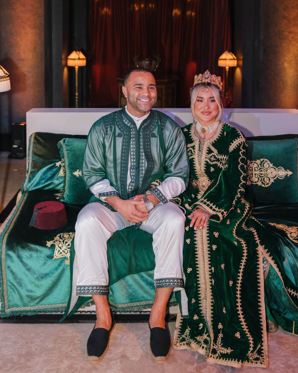 A couple in traditional Moroccan dress sits together on an emerald green velvet couch with ornate gold details.