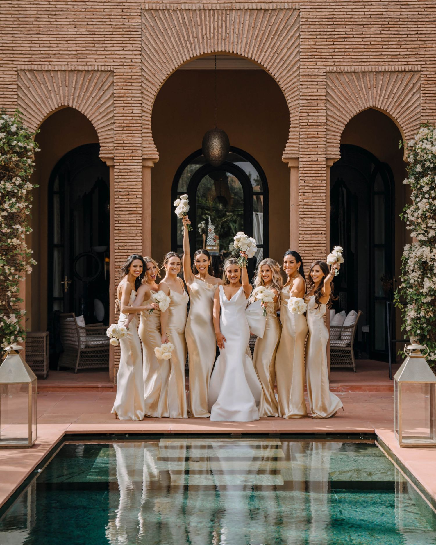 A bridal party in champagne-colored dresses poses by an ornate arched doorway and reflecting pool at a Mediterranean venue.