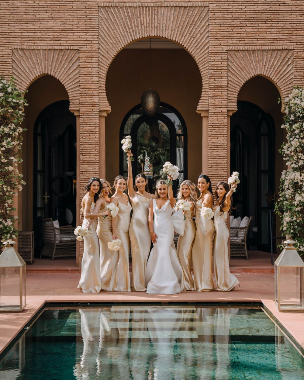 A bridal party in champagne-colored dresses poses by an ornate arched doorway and reflecting pool at a Mediterranean venue.