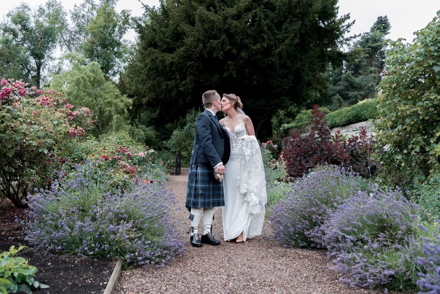 A couple stands on a garden path between blooming lavender and rose bushes in a lush summer garden setting.