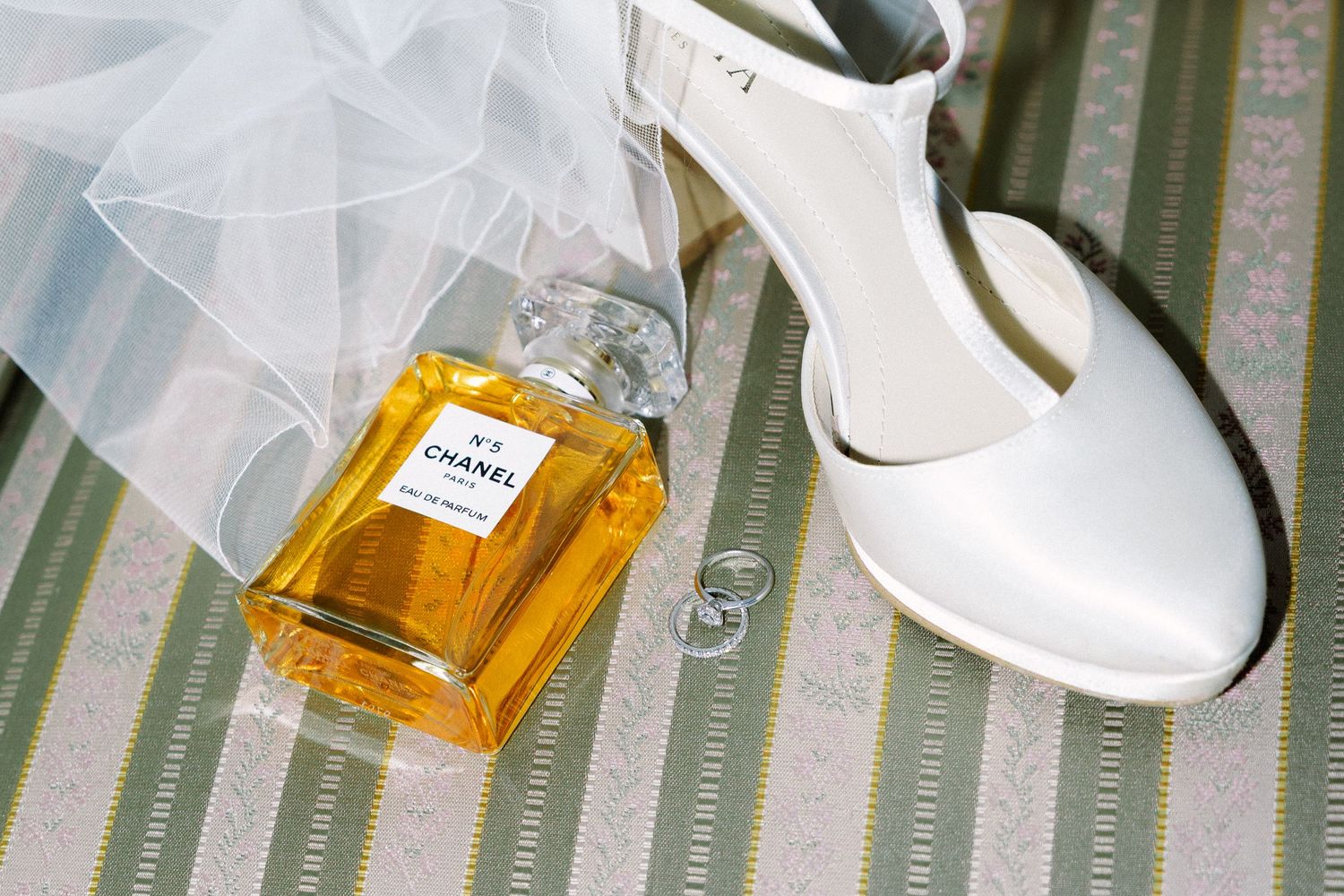 Black and white wedding details featuring white bridal shoes and a Chanel perfume bottle arranged on a striped surface.