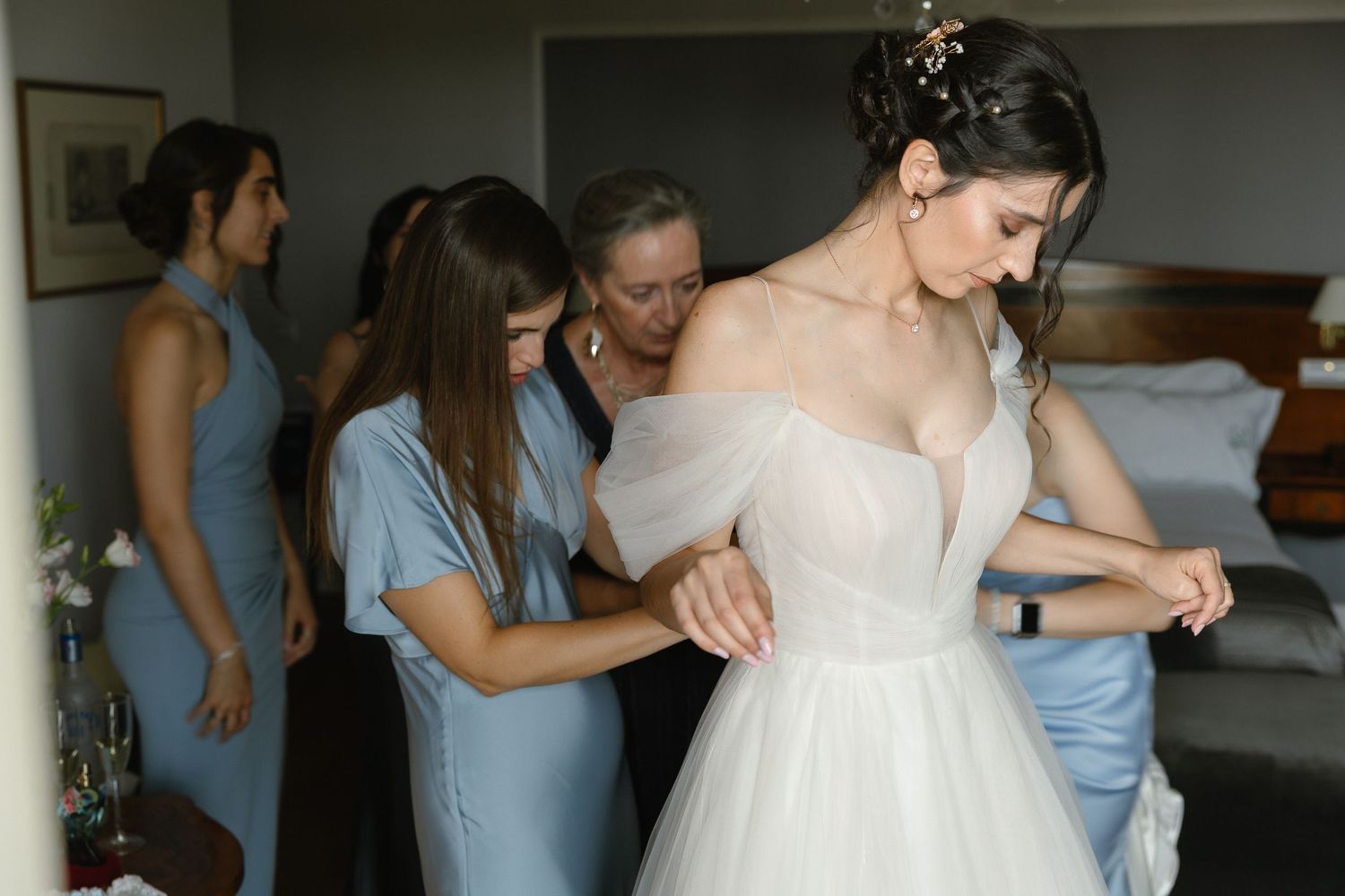 Bridesmaids in light blue dresses help adjust a white wedding gown in a dressing room.