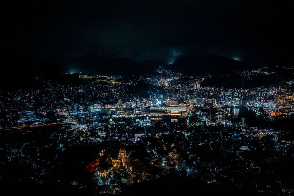 Aerial nighttime cityscape of Nagasaki showing glowing lights and buildings across a dark urban landscape.