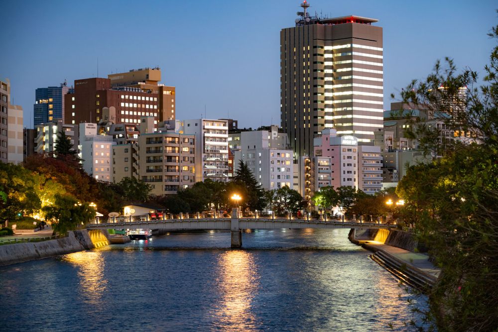 Illuminated waterfront buildings and bridge reflected in calm river waters at dusk.