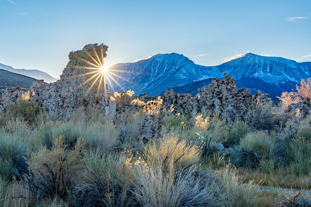 Sunburst through rock formation with mountains and desert brush in foreground.