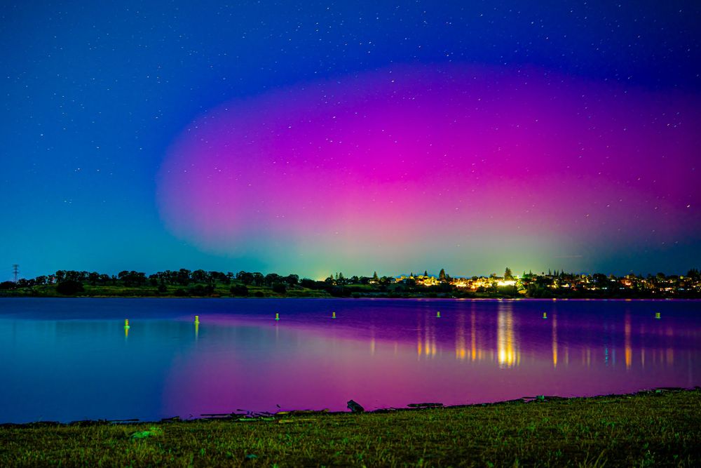 Vibrant purple and blue aurora borealis reflects over calm lake waters at night with city lights in distance.