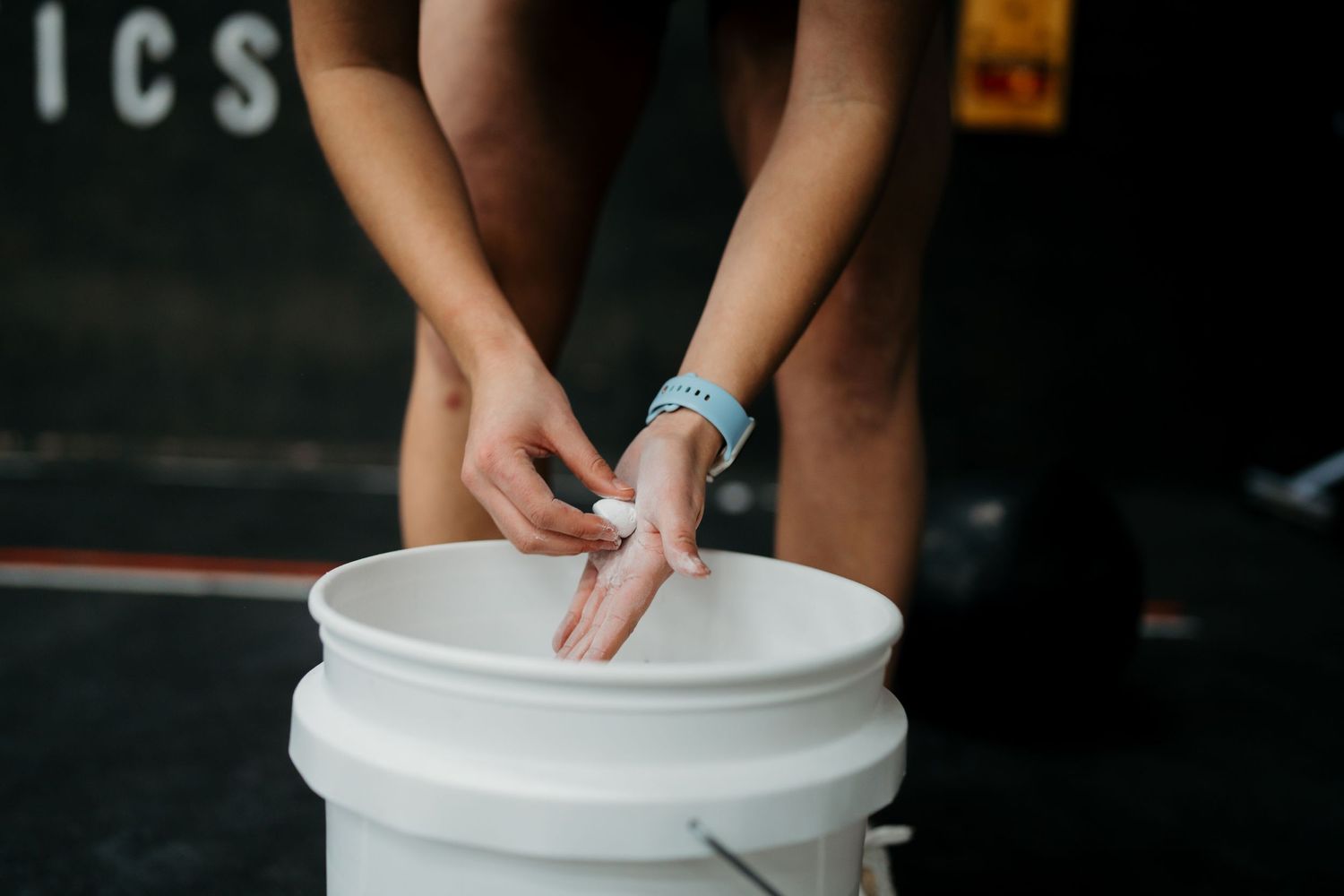 Close-up of hands preparing chalk over a white bucket in a dark gym setting.