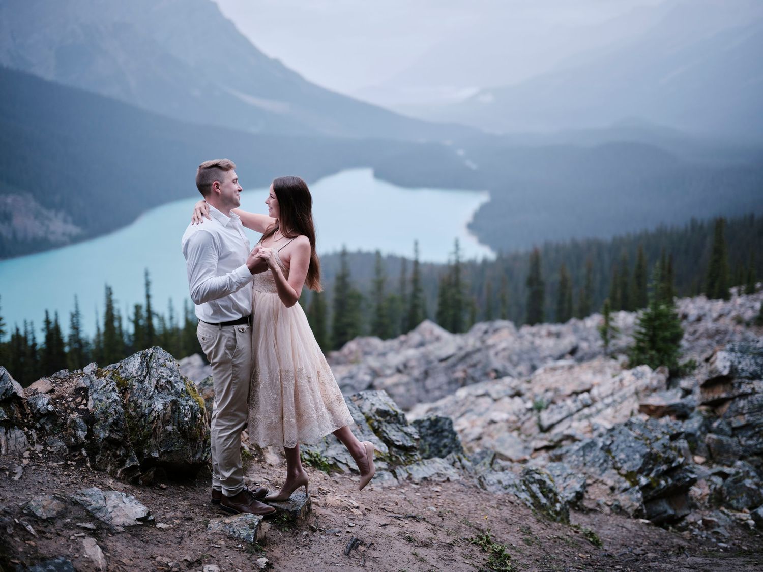 A romantic couple sits on rocky mountain terrain overlooking a misty lake and evergreen forest in the distance. Banff engagement photos