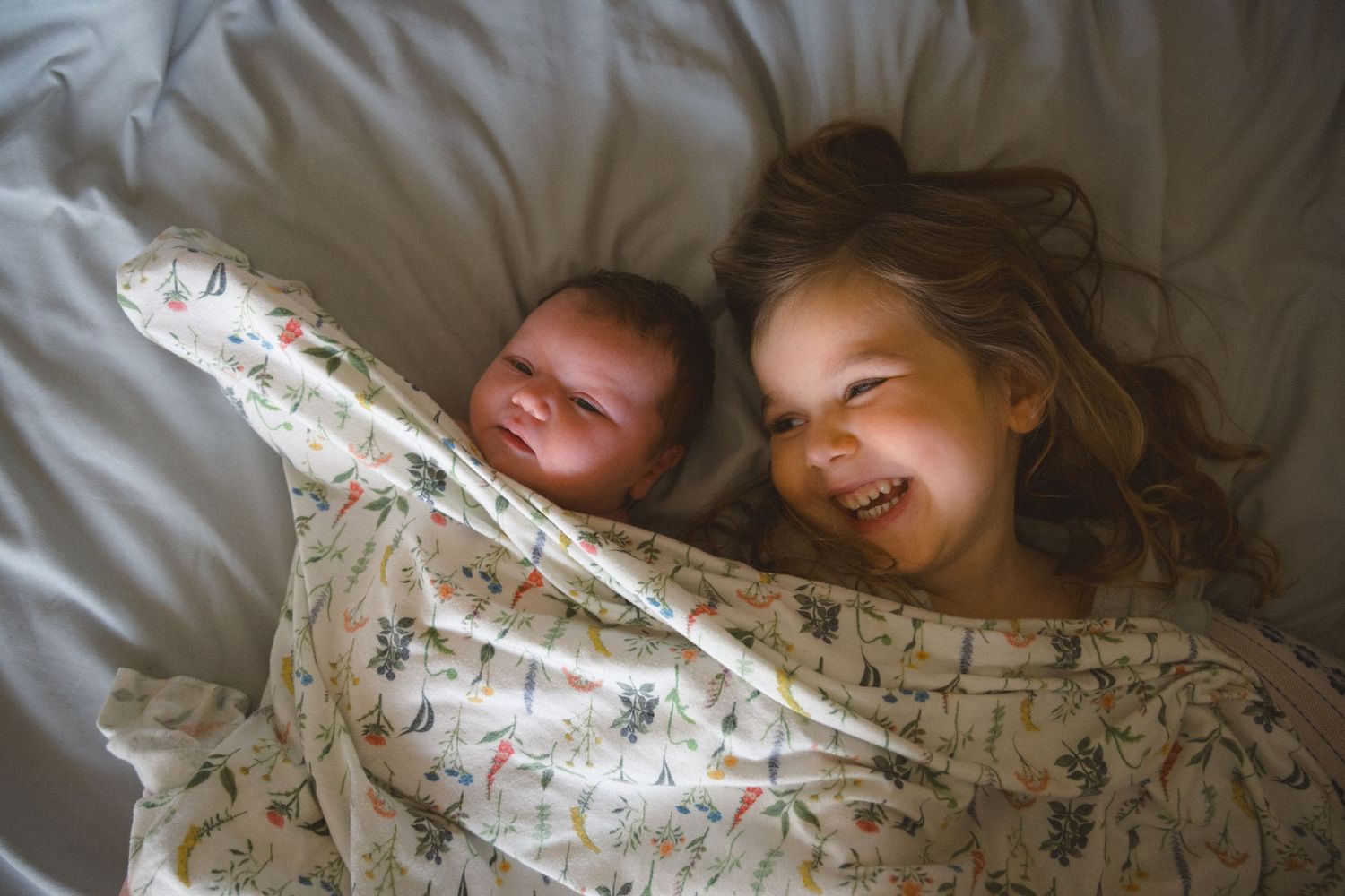 Toddler girl giggling while laying next to newborn baby sister on the bed covered with a floral print baby blanket during newborn photoshoot