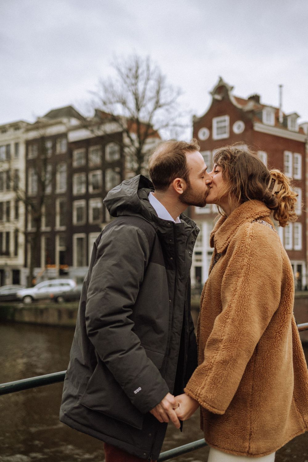 A romantic couple shares a kiss along an Amsterdam canal with historic buildings in the background.