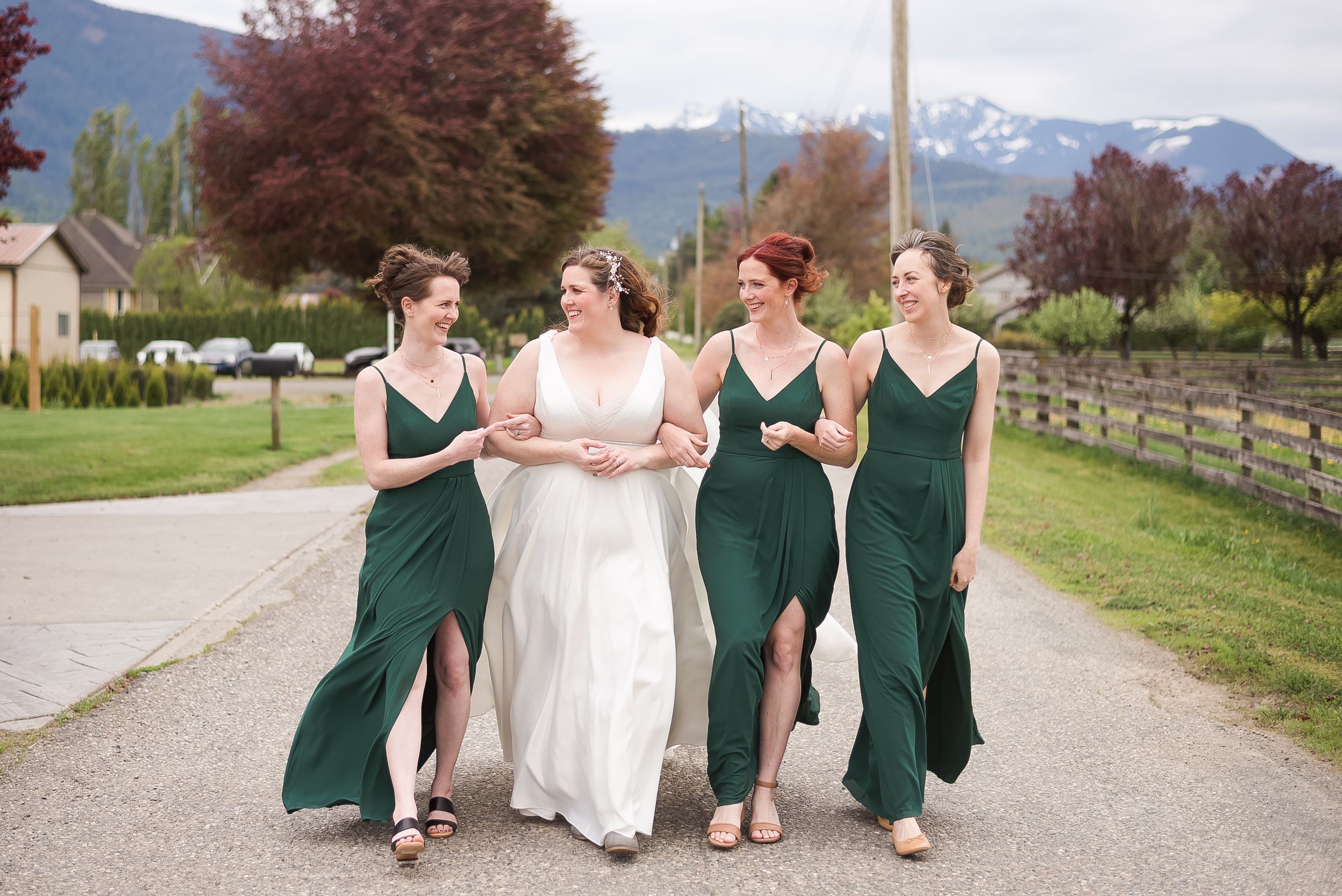 A bride and her bridemaids walk arm in arm with the mountains in the background in Chilliwack, BC.