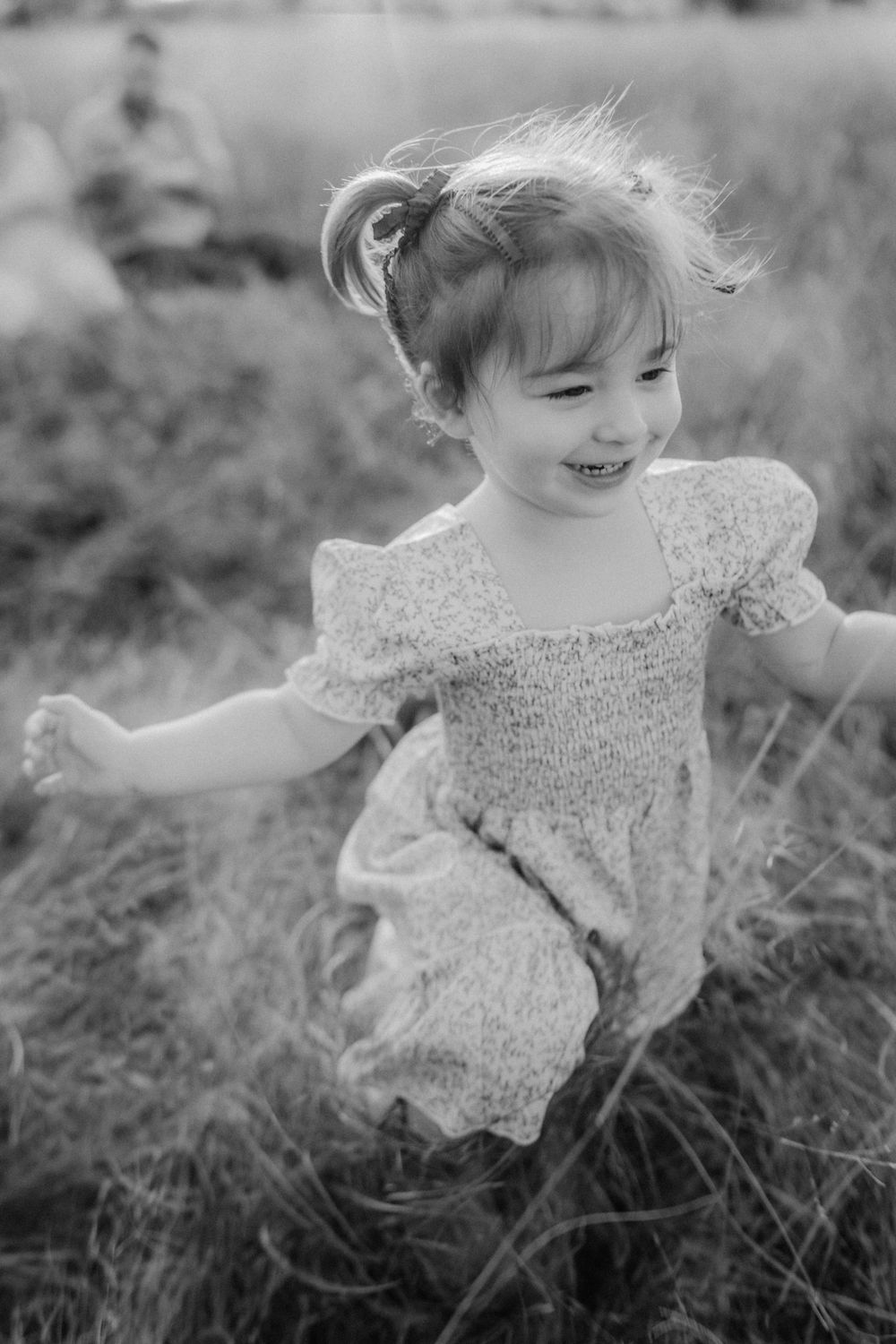 Black and white portrait of joyful child in lace dress playing in tall grass field.