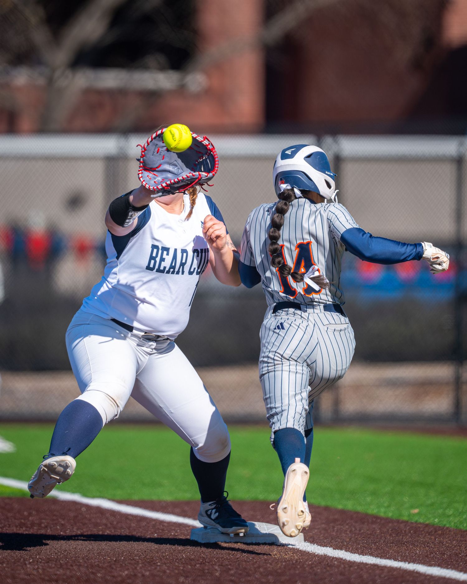 Softball player attempts to catch a yellow ball at first base while runner in pinstriped uniform races to make it safe.
