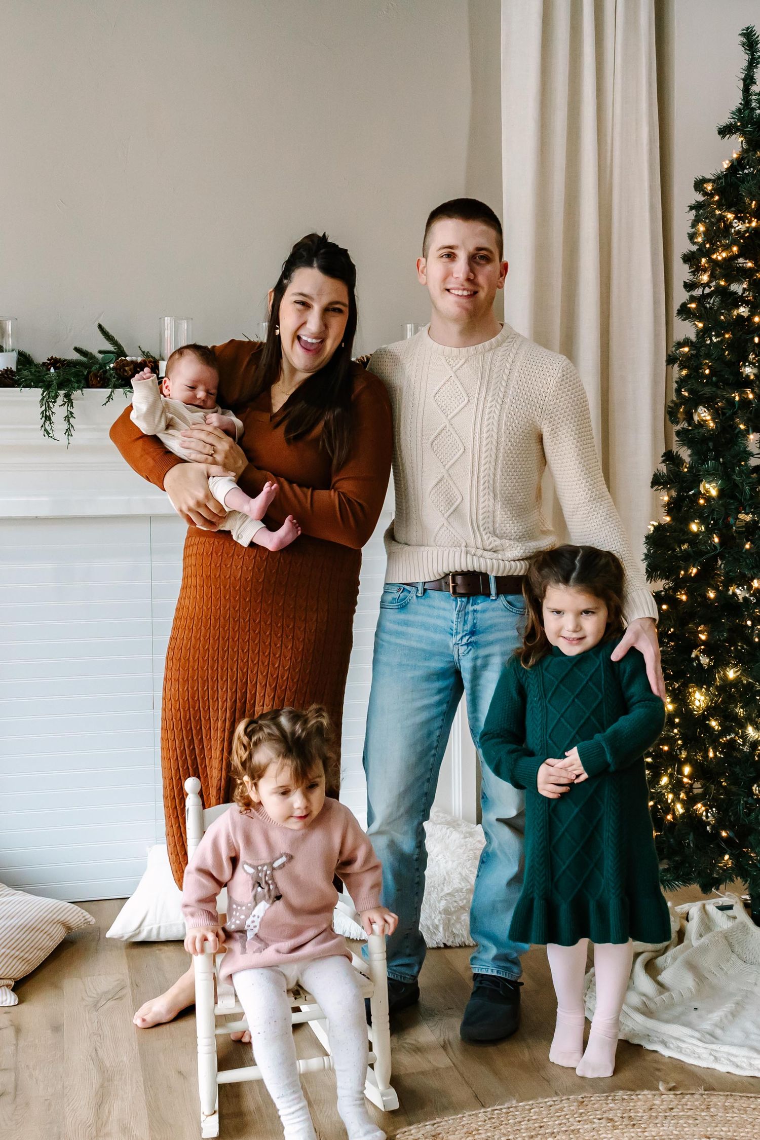 A family poses together in front of a Christmas tree in their cozy living room wearing warm winter sweaters.