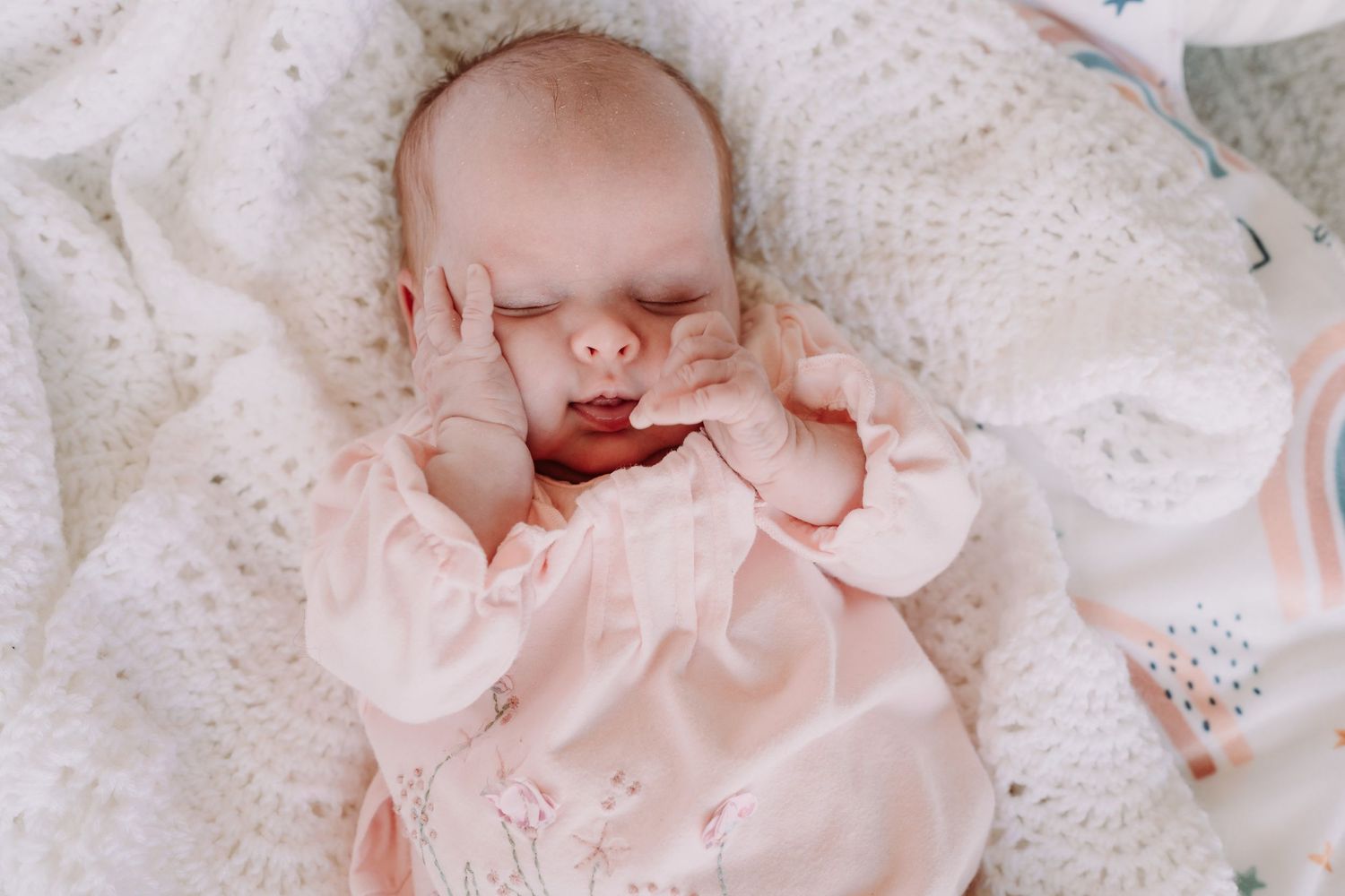 A sleeping infant in a pink onesie rests on a cozy white knit blanket with their tiny hand near their face.