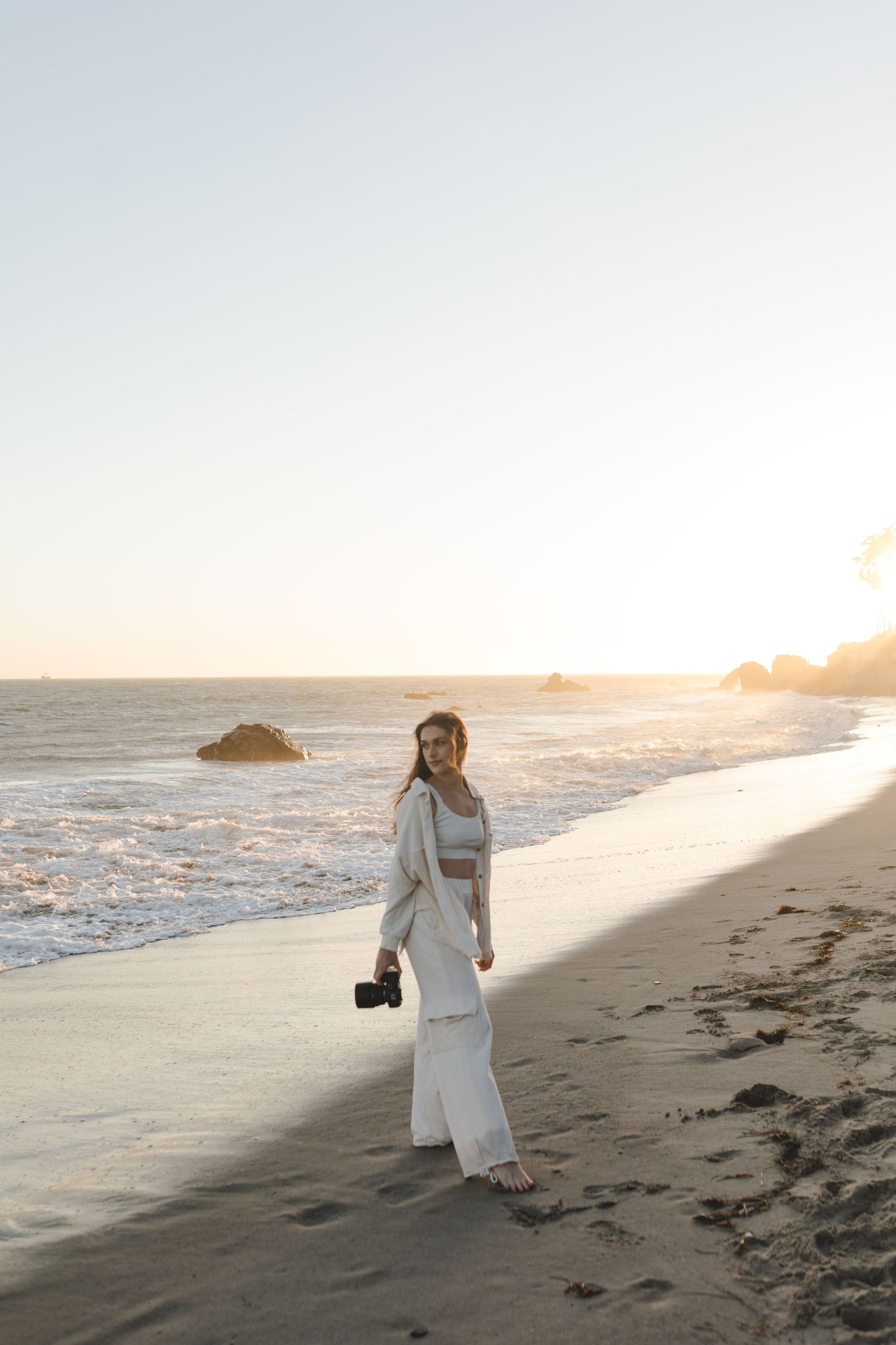 A person in a white outfit stands on a beach at sunset holding a camera while waves crash against rocks in the background.