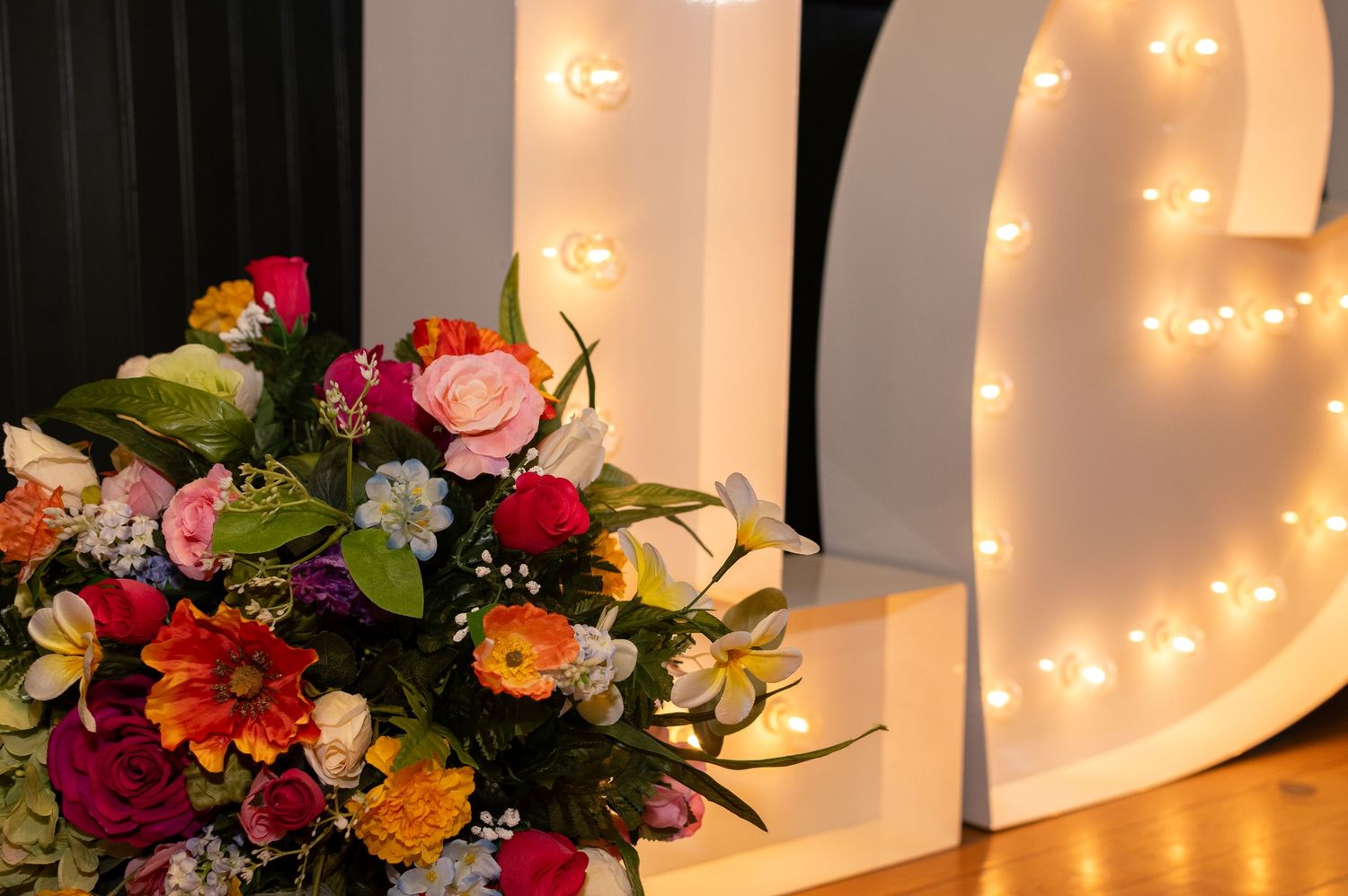A vibrant floral arrangement sits beside an illuminated marquee letter decorated with string lights.